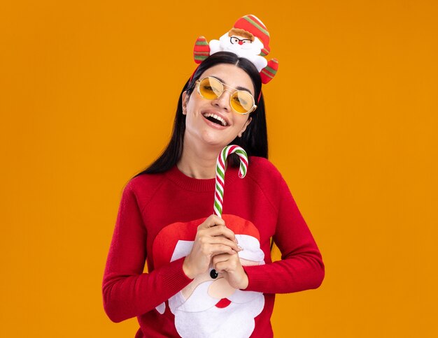 Freudiges junges kaukasisches Mädchen, das Weihnachtsmann-Stirnband und Pullover mit Gläsern trägt, die traditionelle Weihnachtszuckerstange vertikal lokalisiert auf orange Wand mit Kopienraum halten