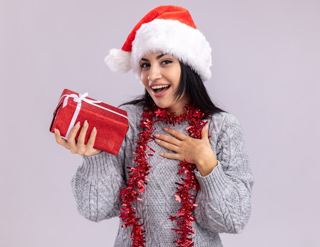 Freudiges junges kaukasisches Mädchen, das Weihnachtsmütze und Lametta-Girlande um den Hals trägt, die Geschenkpaket hält, das Dankeschön lokalisiert auf weißer Wand tut