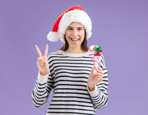 Freudiges junges kaukasisches Mädchen mit Weihnachtsmütze, die Zuckerstange hält und Siegeszeichen gestikuliert