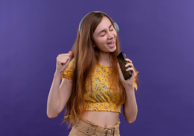 Freudiges junges Mädchen, das Kopfhörer trägt und mit geballter Faust singt, die Handy als Mikrofon auf lokalisiertem lila Raum verwendet