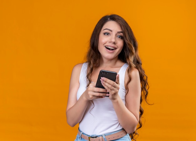Freudiges junges schönes Mädchen, das Handy auf lokalisiertem orange Raum mit Kopienraum hält