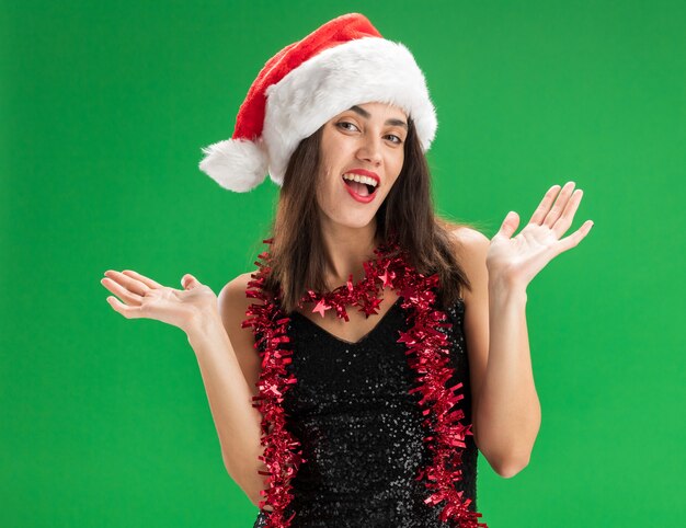 Freudiges junges schönes Mädchen, das Weihnachtsmütze mit Girlande am Hals trägt, die Hände lokalisiert auf grünem Hintergrund spreizt