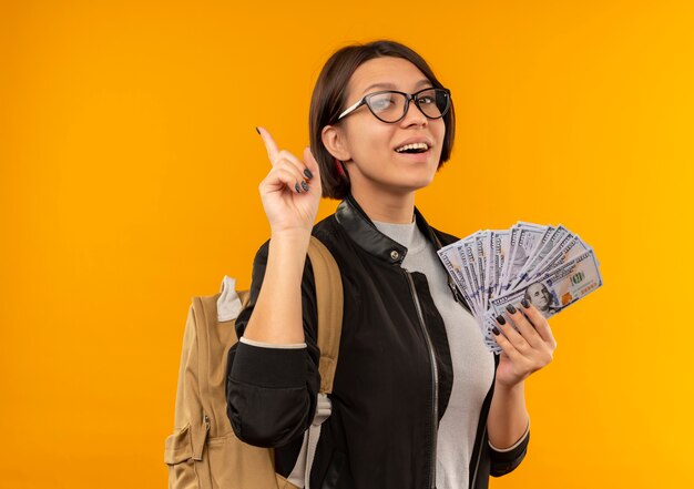 Freudiges junges Studentenmädchen, das Brille und Rückentasche hält, die Geld hält und Finger lokalisiert auf Orange hebt