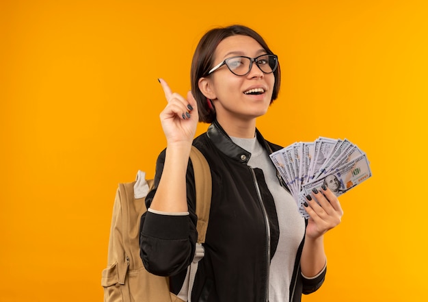 Freudiges junges Studentenmädchen, das Brille und Rückentasche hält, die Geld hält und Finger lokalisiert auf Orange hebt