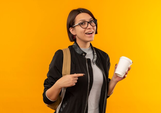 Freudiges junges Studentenmädchen, das Brille und Rückentasche hält und auf Plastikkaffeetasse lokalisiert auf Orange hält