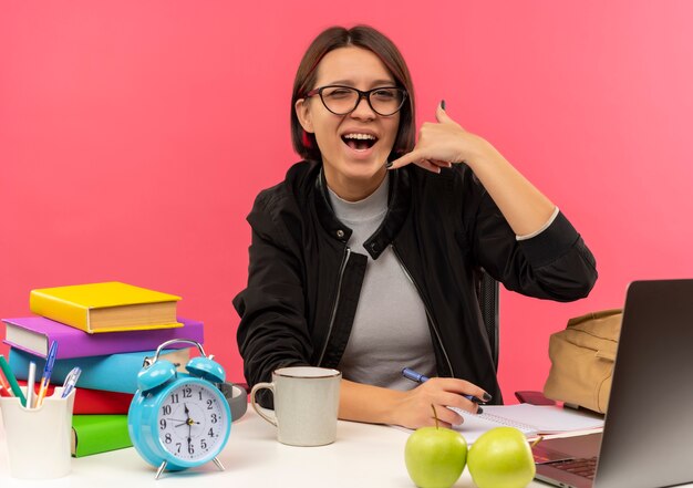 Kostenloses Foto freudiges junges studentenmädchen, das eine brille trägt, die am schreibtisch sitzt und die anrufgeste tut, die hausaufgaben lokalisiert auf rosa tut