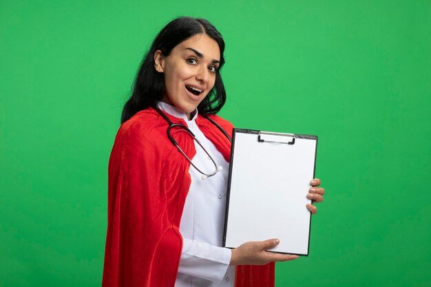 Freudiges junges Superheldenmädchen, das medizinische Robe mit Stethoskop hält Zwischenablage lokalisiert auf Grün trägt