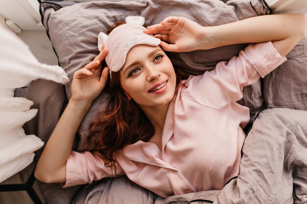Freudiges kaukasisches Mädchen, das am Morgen ruht. Überkopffoto der schönen rothaarigen Frau in der Schlafmaske.