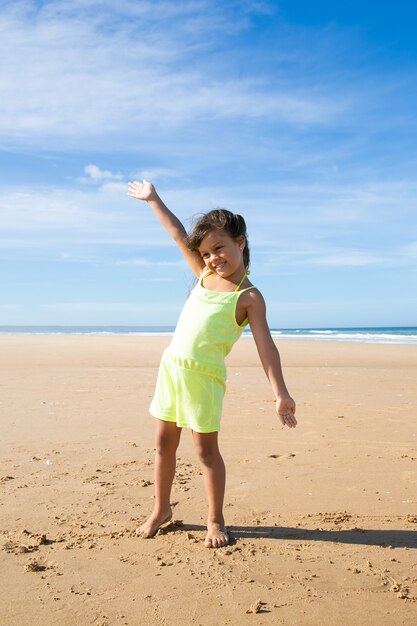 Freudiges kleines Mädchen im Sommertuch, das Aktivitäten am Strand am Meer genießt, mit offenen Armen auf goldenem Sand tanzt und wegschaut