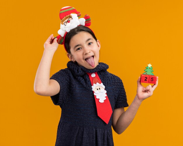 Freudiges kleines Mädchen im Strickkleid, das rote Krawatte mit lustigem Rand auf Kopf hält, der Spielzeugwürfel mit Weihnachtsdatum hält, das Zunge herausragt