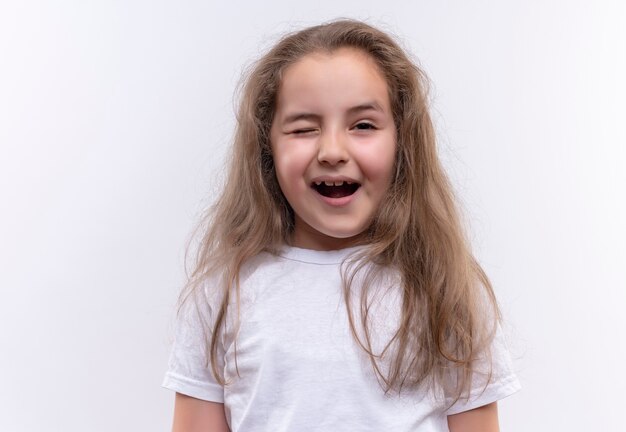 Freudiges kleines Schulmädchen, das weißes T-Shirt trägt, blinzelte auf isolierter weißer Wand