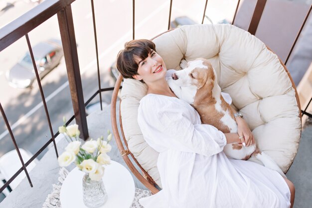 Freudiges Mädchen, das im Stuhl neben Vase der weißen Rosen und des streichelnden Beagle-Hundes sitzt. Schöne braunhaarige Frau, die frische Luft auf Balkon mit Haustierlügen auf ihren Knien genießt