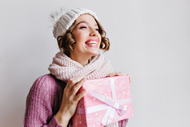 Freudiges Mädchen mit kurzer Frisur, das Winterferien mit Geschenken genießt. Innenporträt der schönen jungen Frau im Pullover und im Schal, die Neujahrsgeschenk auf weißer Wand halten.