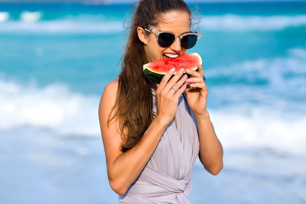 Freudiges Mädchen mit langen hellbraunen Haaren, die eine Wassermelone beißen. Nahaufnahmeporträt des aufgeregten weiblichen Modells in der großen dunklen Sonnenbrille, die Lieblingsessen mit Lächeln genießt.