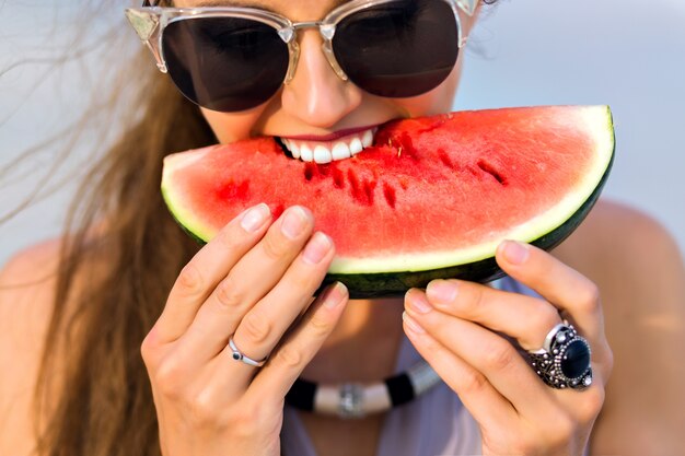 Freudiges Mädchen mit langen hellbraunen Haaren, die eine Wassermelone beißen. Nahaufnahmeporträt des aufgeregten weiblichen Modells in der großen dunklen Sonnenbrille, die Lieblingsessen mit Lächeln genießt.