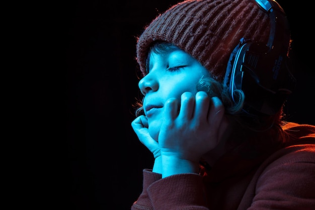 Freudiges Musikhören in Kopfhörern mit geschlossenen Augen. Porträt des kaukasischen Jungen auf dunklem Hintergrund im Neonlicht.