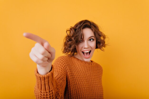 Freudiges romantisches Mädchen mit der trendigen lockigen Frisur, die Finger zeigt und lächelt