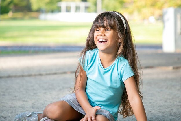 Freudiges süßes schwarzhaariges Mädchen, das auf Boden sitzt, wegschaut und lacht. Konzept für Kindheit und Outdoor-Aktivitäten