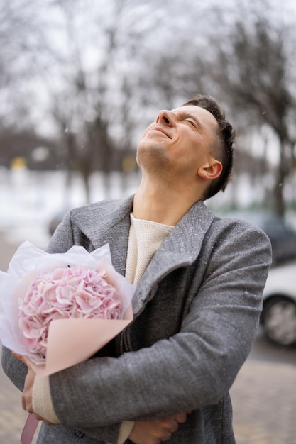 Kostenloses Foto freund mit einem strauß rosafarbener blumenhortensie wartet draußen auf seine freundin, während schnee fällt. valentinstag-konzept, hochzeitsvorschlag. mann geht auf ein date.