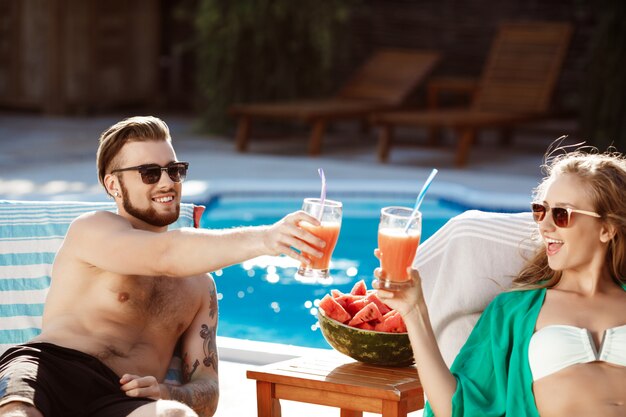 Freunde lächeln, Cocktails trinken, auf Liegen in der Nähe von Schwimmbad liegen