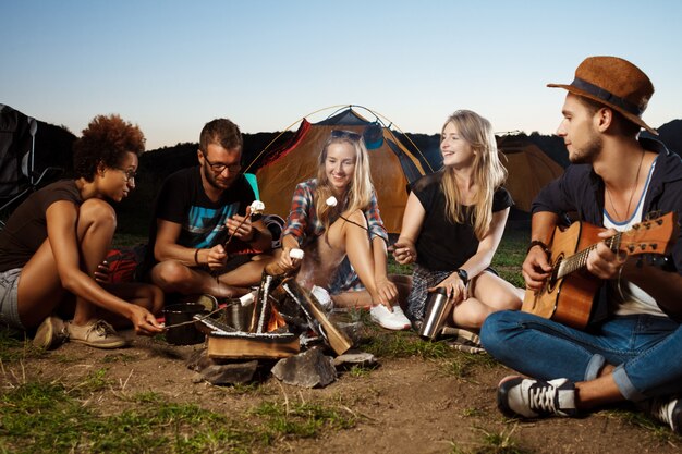 Freunde sitzen in der Nähe von Lagerfeuer, lächeln, Gitarre spielen