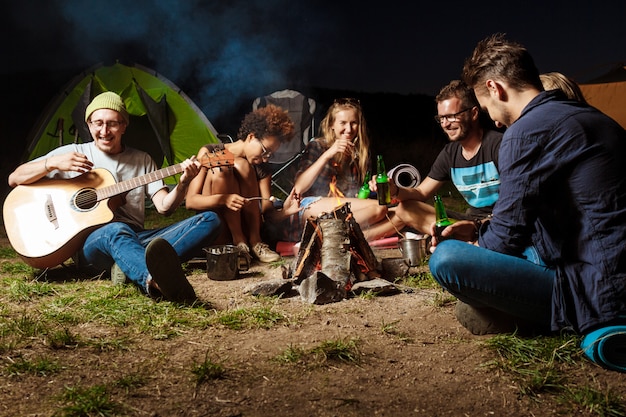 Freunde sitzen in der Nähe von Lagerfeuer, lächeln, sprechen, ruhen sich aus, spielen Gitarre