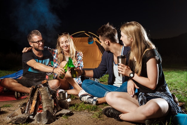 Freunde sitzen in der Nähe von Lagerfeuer, lächeln, sprechen, ruhen sich aus, trinken Bären