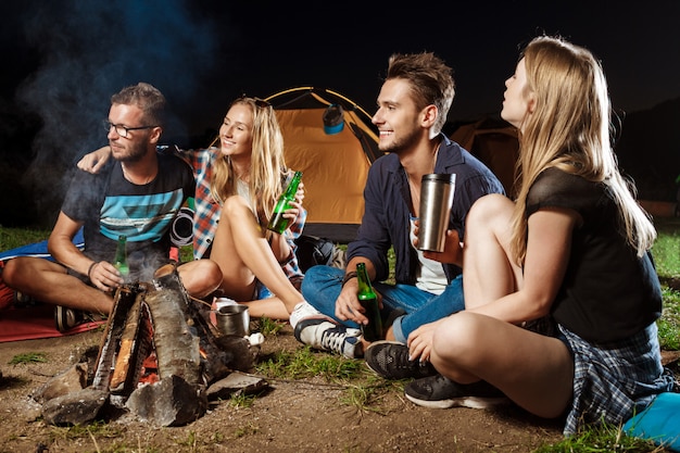 Freunde sitzen in der Nähe von Lagerfeuer, lächeln, sprechen, ruhen sich aus, trinken Bären