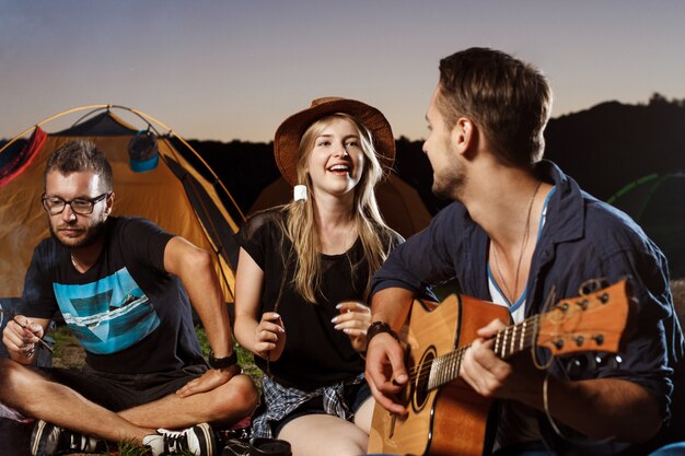 Freunde sitzen in der Nähe von Lagerfeuer, lächelnd, spielen Gitarre Camping Grill Marshmallow.