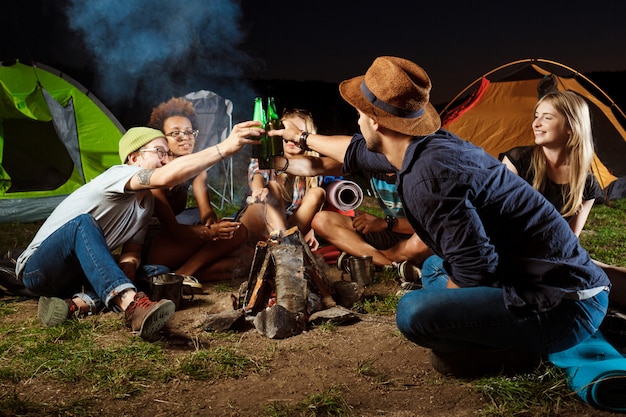 Freunde sitzen in der Nähe von Lagerfeuer, trinken Bären, lächeln, sprechen, ruhen