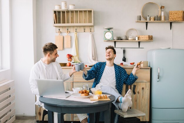 Freunde, welche die Kaffeetasse sitzt mit neuem Lebensmittel und Laptop auf Tabelle zujubeln