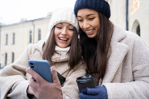 Freundinnen, die nach der wiedervereinigung auf das smartphone schauen und kaffeetassen im freien halten