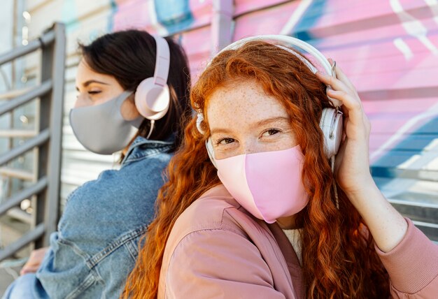 Freundinnen mit Gesichtsmasken im Freien hören Musik auf Kopfhörern