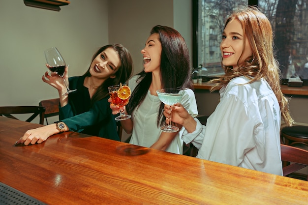 Freundinnen trinken an der Bar. Sie sitzen an einem Holztisch mit Cocktails. Sie stoßen an