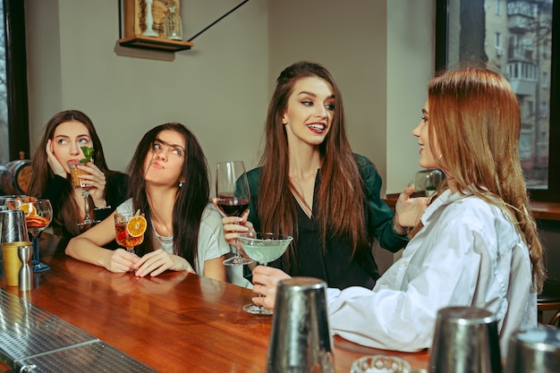 Freundinnen trinken an der Bar. Sie sitzen an einem Holztisch mit Cocktails. Sie tragen Freizeitkleidung.