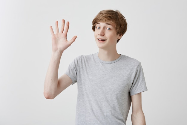 Freundlich aussehender höflicher junger kaukasischer Mann gekleidet in grauem T-Shirt lächelnd, hallo sagend, seine Hand winkend. Positive menschliche Emotionen, Mimik, Gefühle, Haltung und Reaktion