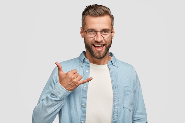 Freundlich aussehender positiver Hipster mit dunklen Stoppeln, Gesten im Innenbereich, zeigt Shaka-Zeichen, in Hochstimmung