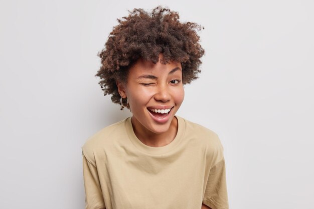 Freundliche fröhliche Frau hat verspielte Stimmung zwinkert Auge lächelt breit gekleidet in lässiges beiges T-Shirt gekleidet in lässiges braunes T-Shirt isoliert über weißem Hintergrund Positives Konzept menschlicher Emotionen