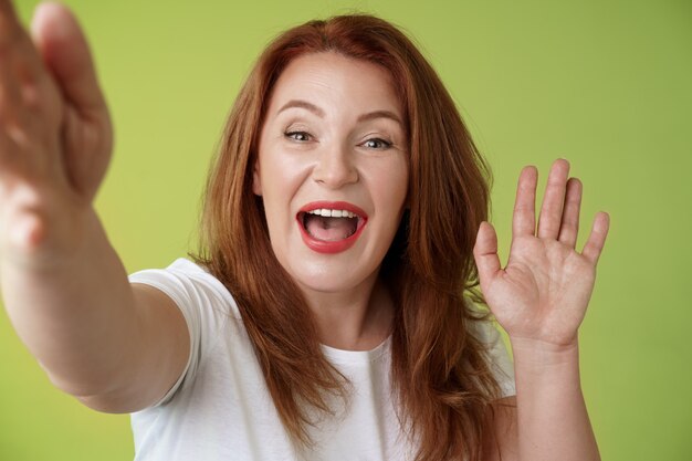 Freundliche fröhliche rothaarige Frau mittleren Alters verlängern Arm halten Kamera unter Selfie winkende Handfläche Hallo Hallo Gruß lächelnd breit willkommen Tochter sprechen Videocall Mobile Internet grüne Wand