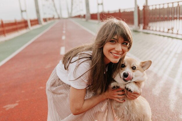 Freundliche, glückliche Kaukasierin mit rosa Rock und weißem T-Shirt, die mit ihrem kleinen süßen Hund in die Kamera posiert