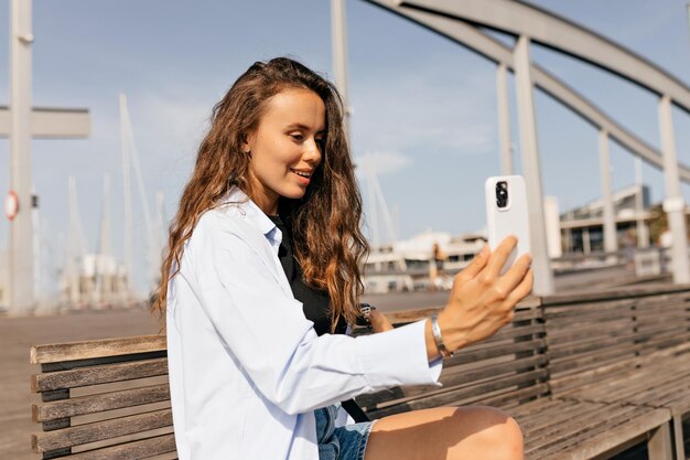 Freundliche junge europäische lächelnde Frau winkt mit der Hand auf den Smartphone-Bildschirm, der an sonnigen Tagen im Freien ist Langhaarige Brünette trägt blaues Hemd und Hemden, die auf dem Pier sitzen Telefonkommunikationskonzept
