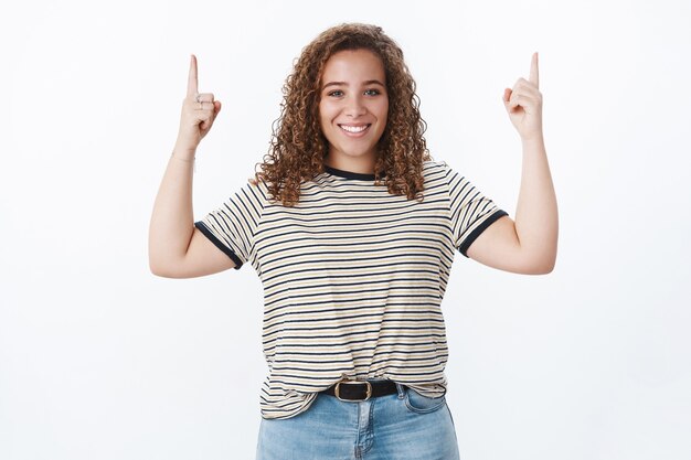 Freundliche, lebhafte, energiegeladene, charmante junge Frau, weiße Zähne, die die Richtung zeigen, die nach oben zeigen, erhobene Zeigefinger, die eine coole Promo fördern und Unsicherheiten bekämpfen, heben das Selbstwertgefühl nach oben