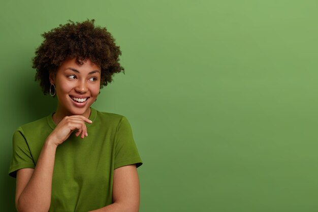 Freundliche optimistische junge afroamerikanische Frau schaut zur Seite, hält die Hand sanft unter dem Kinn, stellt sich ein großartiges Ereignis vor, trägt ein lässiges grünes T-Shirt, posiert im Innenbereich und bietet Platz für Ihre Werbung