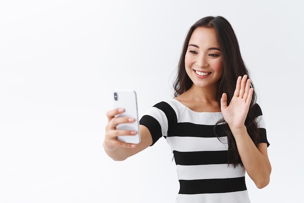 Freundliche, zarte junge Asiatin in lässigem Outfit, die über Internet-Videoanruf spricht und ein Smartphone hält, das ein Selfie mit der Hand winkt, in einer Hallo-Grußgeste, die auf das mobile Display lächelt