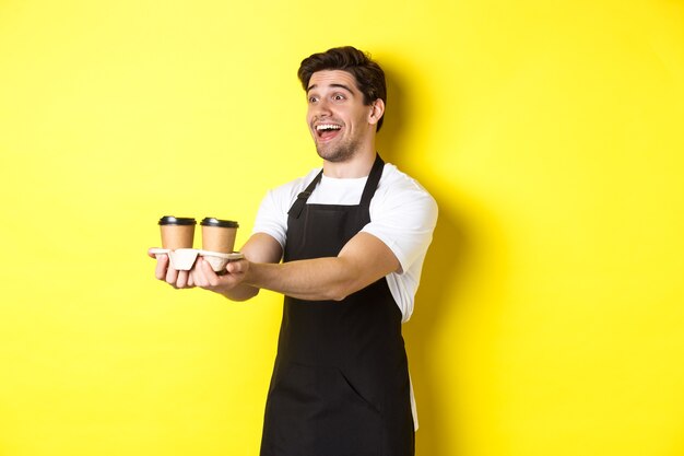 Freundlicher Barista in der schwarzen Schürze, die Bestellung zum Mitnehmen gibt, zwei Tassen Kaffee hält und lächelt und über gelbem Hintergrund steht.