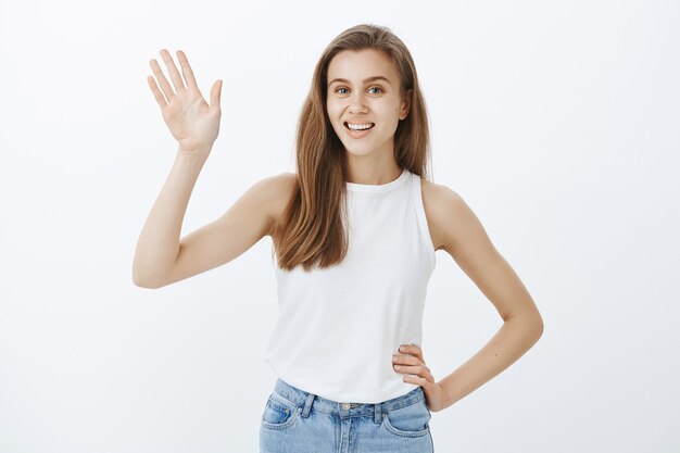 Freundliches blondes Mädchen, das lächelt und Hand winkt, um Hallo zu sagen, jemanden zu begrüßen, hallo Geste