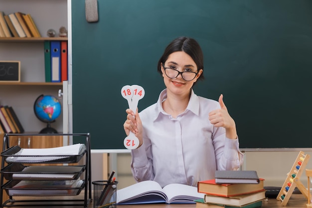 freut sich, Daumen hoch zu zeigen junge Lehrerin mit Brille, die Spaß am Schreibtisch mit Schulwerkzeugen im Klassenzimmer hält