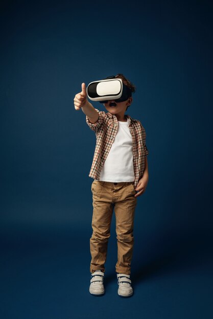 Frieden für andere Planeten. Kleiner Junge oder Kind in Jeans und Hemd mit Virtual-Reality-Headset-Brille einzeln auf blauem Studiohintergrund. Konzept der Spitzentechnologie, Videospiele, Innovation.