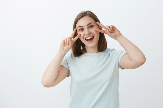 Friedliche glückliche und freundliche gut aussehende Frau im trendigen T-Shirt, das Friedens- oder Siegeszeichen über Gesicht zeigt und breit lächelnd positive und fröhliche Schwingungen ausdrückt, die amüsiert über weißer Wand stehen