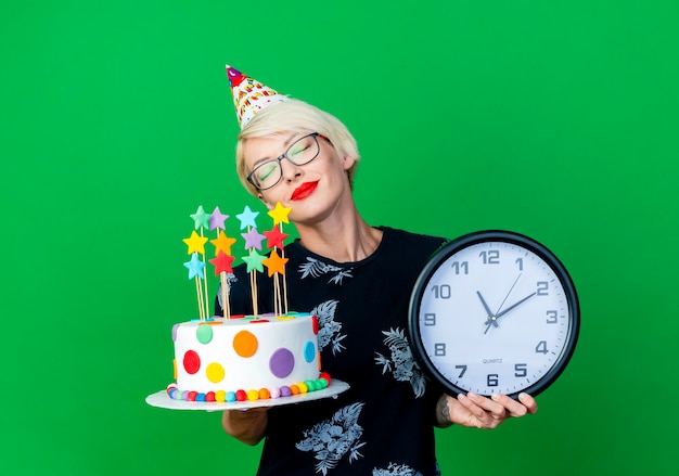 Friedliche junge blonde Partygirl, die Brille und Geburtstagskappe hält, die Geburtstagstorte und Uhr mit geschlossenen Augen lokalisiert auf grünem Hintergrund mit Kopienraum hält